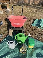 Full lot setup showing red EarthWay spreader, two watering cans in green and grey, green plant food container, and bird house on a green tarp
