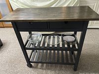 Front view of kitchen island showing 2 drawers, slatted lower shelves with glass dishes inside, and wheels for mobility.