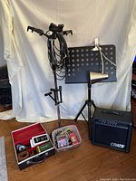 Wide shot of the entire lot showing the multi-instrument stand with cables, music book stand with clip-on light, Crate GX15 amplifier, and two cases with spare parts on wooden floor against white background.