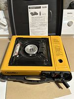 Front view of portable butane gas cooker with yellow casing, black burner, control knob, and instruction manual inside the lid.