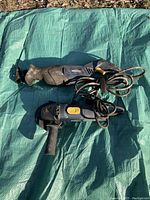 Two power tools lying on a green tarp outdoors; one is a reciprocating saw with a blade and the other is an angle grinder with a side handle and cord.