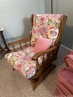 Front perspective of the rocking chair showing floral padded cushions, wooden armrests with spindles, and pink accent pillow.