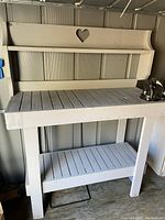 Front angle of white wooden work bench showing slatted shelf and decorative backboard with heart cutout. Attached black metal vise mounted on right side of bench.