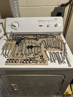 Photo showing a variety of chrome-finished open-end and combination wrenches, socket wrench handles, and various sockets laid out on top of a washer/dryer appliance.