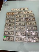 Full display of 23 two dollar bills laid out on a glass table, some protected in plastic sleeves, showing the front design featuring Thomas Jefferson.