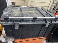 Front and side view of a large black rectangular DeWalt rolling tool box with wheels and metal handle bars, showing signs of heavy use and dirt.