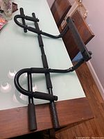 Pull up bar laid flat on wooden dining table, showing multiple handles with foam grips and black metal frame.