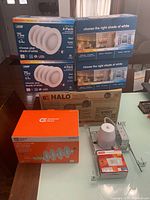 Photo showing multiple boxed LED light fixtures and trims on table, including Commercial Electric and Halo brands