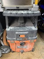 Front view of Ridgid wet/dry vacuum with on/off switch, hose inlet, and Ridgid logo visible. Dust on surface.