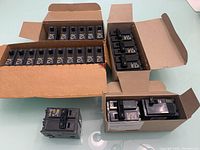 Multiple cardboard boxes containing black Square D circuit breakers with toggle switches; most breakers marked 20 amps.