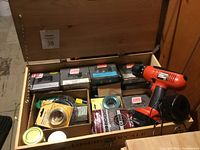 Top view of wooden box with drill, boxes of screws, wires, steel wool, and other hardware items inside.