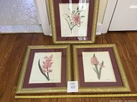 Three framed floral prints displayed together on the floor, showing the full look of the soft gold frames with maroon and gold mats and pink flower illustrations.