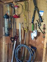 Full wall shot showing various bird feeders, garden tools, spades, bungee cords, and hooks mounted on pegboard and wood wall.