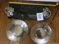 View of green marble rectangular cheese tray with metal grape leaf and berry shaped handles and two small glass plates on wooden surface with measuring tape.