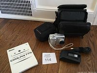 Nikon CoolPix 775 digital camera with user manual, carrying cases, and battery charger on wooden floor
