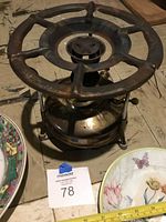 Optimus Number 45 camp stove with rusted cast iron top, metal fuel tank, showing wear. Several ceramic serving plates partially visible around it.