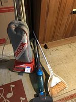 Oreck upright vacuum, blue lightweight Bissell vacuum, and white broom and dustpan on floor against wood panel wall.