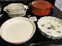 Lot of five cookware and bakeware items displayed on stove including two pastry tart dishes, Corning Ware casserole dish with glass lid, orange Dansk metal pot with lid, and white ceramic dish with vegetable decoration.