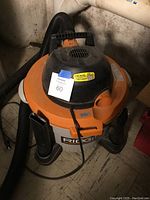 Shop vacuum front view showing Rigid branding, orange top, gray canister, black motor housing and connected hose.