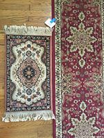 Both small runner rugs laid side by side on a wood floor. Larger rug is maroon and tan with floral geometric border. Smaller rug is salmon, navy, cream, and tan with tassels.