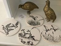 Four black and white ceramic waterfowl trivets and two brass partridge figurines displayed together