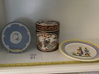 Three items shown on shelf: Wedgwood blue jasperware decorative plate, Henriot Quimper porcelain canister, hand-decorated Macau plate