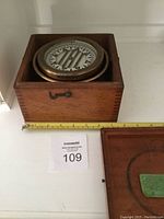 Compass housed in wooden box showing front and top view with measuring tape for scale, keys and latch visible