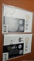 Two GRA certification cards for the moissanite earrings, displaying carat size 2.0ct each, 8.0mm, D color, VVS1 clarity, with warranty ID codes and QR codes.