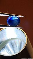 Close-up photo showing an oval faceted natural blue sapphire gemstone held by tweezers above a loupe magnifier. The stone shows bicolor dark and medium blue color.