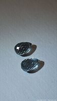 Two light blue, pear-shaped aquamarine gemstones, cut in fancy briolette style showing faceted surfaces and clarity, placed side by side on neutral background.