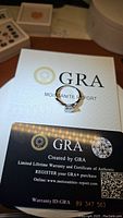 Photo showing the ring on top of a GRA Moissanite Report card and a warranty certificate card with barcode and warranty ID, indicating authenticity and certification.