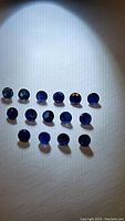 Fifteen natural deep blue diamond cut sapphires arranged on a white surface under light, showing size and color.
