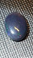 Close-up photo of an oval black opal cabochon showing multicolored flashes under bright light on textured background.