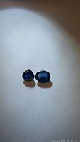 Pair of natural blue sapphire gemstones on neutral background showing oval and pear cuts with rich blue color and clarity.