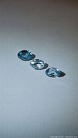 Three oval cut natural Cambodian zircon gemstones, light blue and colorless tones, on white background showing clarity and cut details.