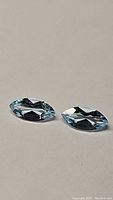 Pair of marquis cut natural blue topaz gemstones placed side by side on a flat surface, showing their elongated shape, faceted surface and light blue color.