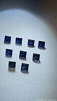 Eight natural blue sapphires cut in princess shape arranged on a white background under bright lighting.