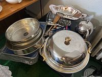 Photo showing large metal covered serving dish, round baking pans, metal trays, teapot, and assorted cutlery, placed on green carpeted floor near wall corner.