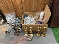 Overview of the lot showing one silver 5-arm metal candelabra, three pairs of brass candlestick holders, a small decorative brass candle holder, bags of white tea light candles, pillar candles and assorted small holders inside cardboard box.