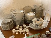Wide view showing complete assortment of porcelain dishes on table including cups, plates, bowls, teapot, egg cups, and serving pieces with purple floral pattern and gold trim.