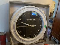 Large round wall clock with white frame and black face still in damaged packaging, showing front view.