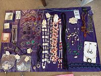 Overview of assorted costume jewelry items spread on a purple cloth including necklaces, bracelets, charms, earrings, and pendants in packaging or loose.