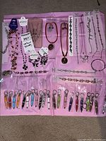 Full overview of costume jewellery lot with necklaces, bracelets, and keychains on a pink background. Items mostly in original packaging, showing variety of styles and colors