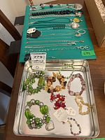Photo showing bracelets displayed on a metal tray with assorted styles and colors, plus silver-tone chain necklaces on a green board.