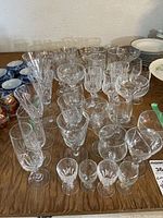 Wide shot showing assortment of glass and crystal glassware on wood table, including tall champagne flutes, wine glasses and small cordial glasses.