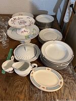 Photo capturing variety of dishes including cake stands, stacks of white plates and soup bowls, serving platters, tureen, creamer, and sugar bowl on wooden surface.