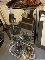 Wide shot showing the beveled wall mirror leaning on the floor with silver plate tea set arranged on the tray placed in front and decorative metal trays hanging above the mirror.