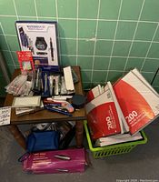 Photo showing Waterman fountain pen set, various pens, keychains on small wooden table beside boxes of ruled paper in a green basket.