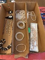 Boxes containing various pearl bracelets and necklaces in different colors including white, pink, purple, and black bead accents