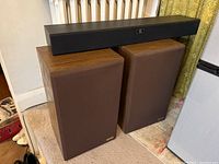 Two Fidelio RS200 rectangular speakers with wood finish and brown fabric covers, with one long black wireless speaker on top.